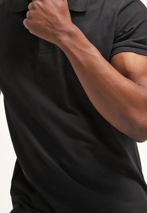 Personne portant un polo noir à manches courtes, la main droite reposant près de l'épaule gauche.