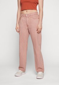 Jean en denim rose clair à taille haute, jambe droite, avec des détails de couture visibles. Associés à des baskets blanches et un top court de couleur rouille.