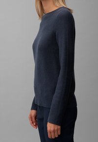 Kobieta w dopasowanym, ciemnoniebieskim swetrze z teksturowanym splotem z długimi rękawami oraz granatowych spodniach, na tle jednolitego szarego tła.