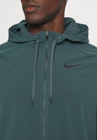 Chaqueta con capucha de Nike en verde oscuro, con cierre de cremallera completo, cordones y un logo negro. Ligera, de textura suave y con costuras mínimas.