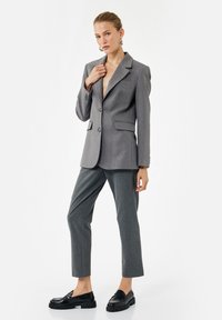 Gray tailored blazer with sharp lapels, two buttons, and side pockets, paired with gray trousers and glossy black loafers. Smooth fabric texture.