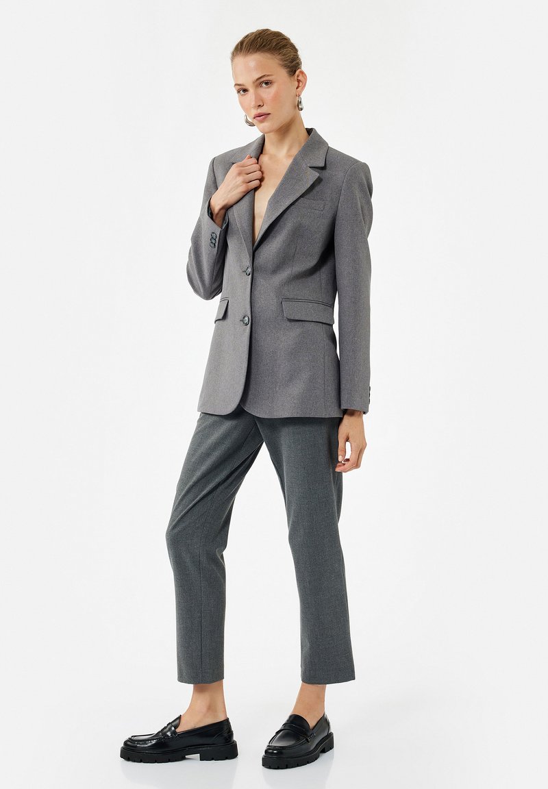 Gray tailored blazer with sharp lapels, two buttons, and side pockets, paired with gray trousers and glossy black loafers. Smooth fabric texture.