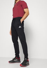 Man i svarta Adidas-joggers, svarta och röda Adidas-sneakers och en vinröd Adidas-t-shirt, stående med händerna i fickorna.