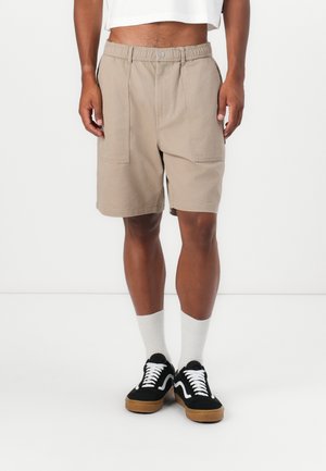 Pantalones cortos beige con cinturilla elástica, dos bolsillos frontales y un ajuste relajado, combinados con zapatillas negras y calcetines blancos de caña media.