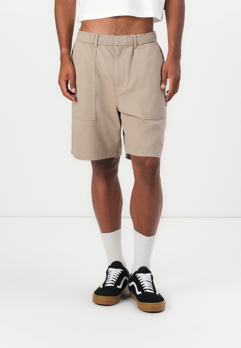Beige shorts med elastisk midja, två framfickor och avslappnad passform, tillsammans med svarta sneakers och vita ankelsockor.