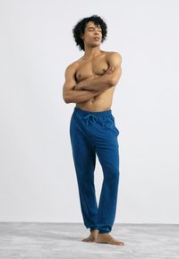 Blauwe drawstring broeken met een ontspannen pasvorm, met een soepele textuur. Persoon staat blootsvoets met de armen gekruist tegen een effen achtergrond.