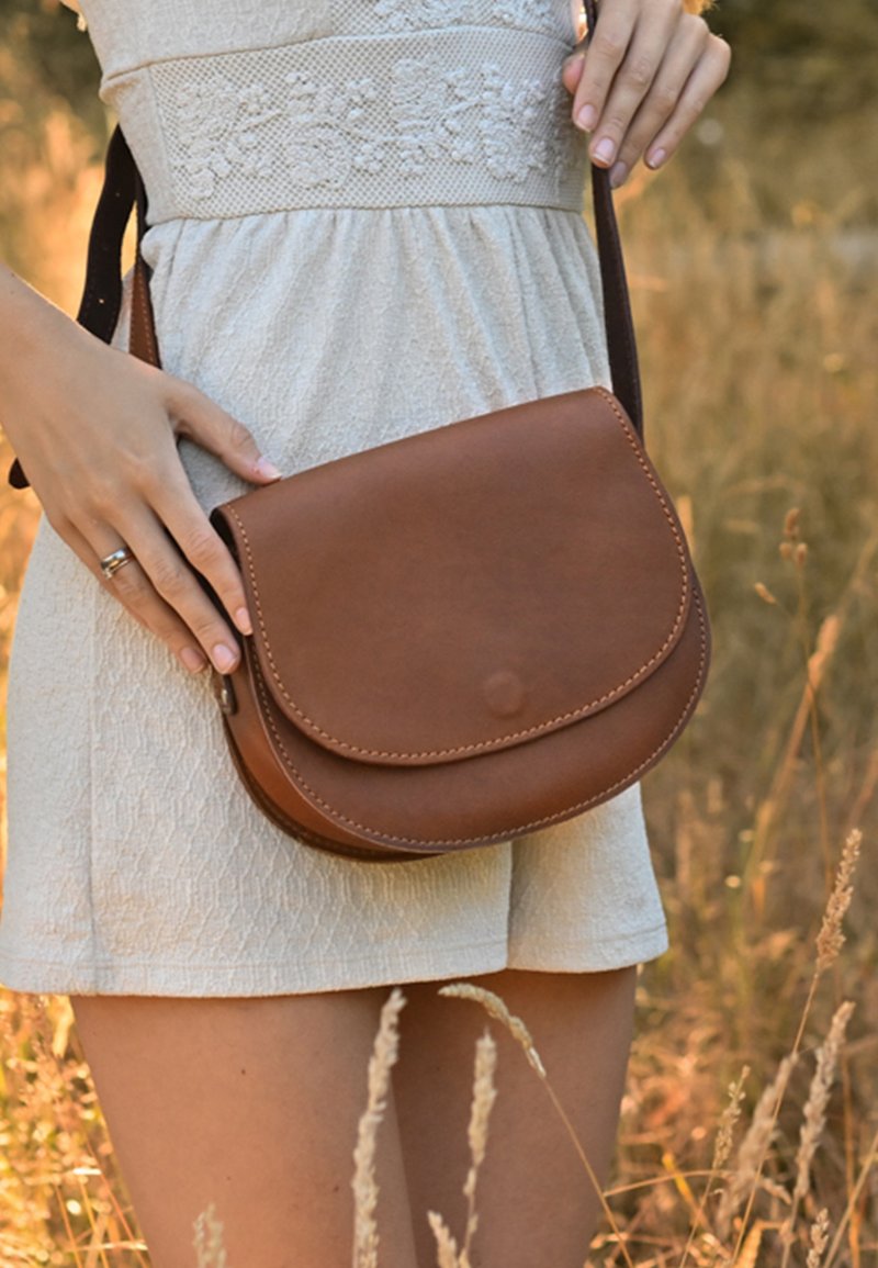 Brązowa skórzana torebka na pasku z zaokrągloną klapką, przeszytymi krawędziami i gładką fakturą, trzymana przez osobę na tle naturalnego krajobrazu.