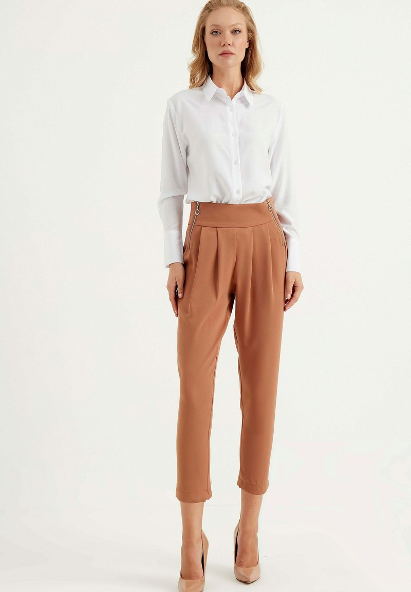 Witte button-up shirt gecombineerd met hoog-waist bruine broeken met zijritsen en plooien, afgewerkt met nude hoge hakken.