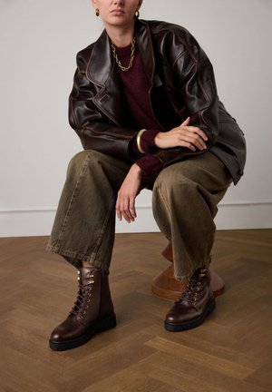 Brown leather ankle boots with gold eyelets, black rubber sole, textured surface, worn with olive green wide-leg pants and a dark sweater.