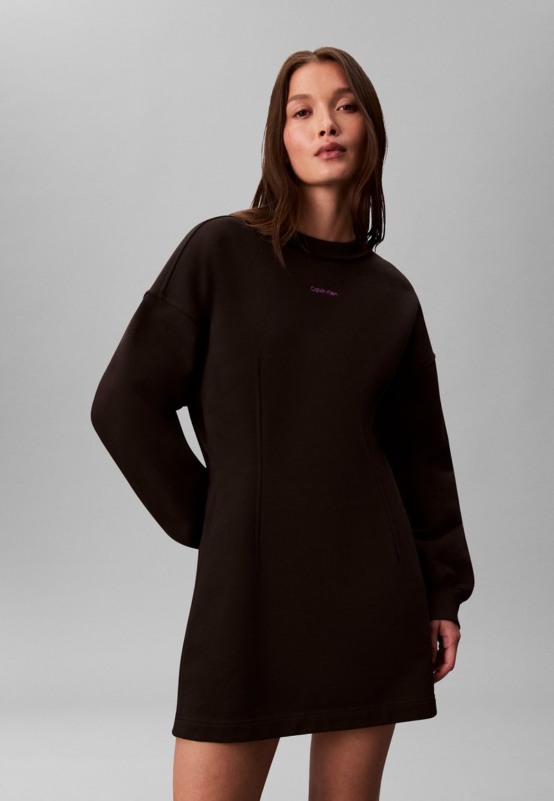 Robe sweat-shirt noire, design oversize, col rond côtelé et manches longues. Présente un petit logo violet sur la poitrine. Texture lisse.