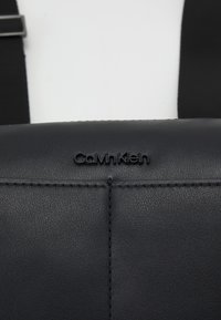 Črna usnjena torba za rame z vidnimi šivi, embosiranim logotipom Calvin Klein in nastavljivim tekstilnim trakom. Splošna gladka tekstura.