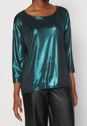 Femme portant un haut à manches longues bleu sarcelle brillant avec un col rond et un pantalon noir aspect cuir sur un fond uni.