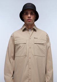 Camisa de botones beige con dos bolsillos en el pecho, fabricada en un tejido suave y con un corte relajado, combinada con un sombrero de cubo negro que presenta un logo circular.