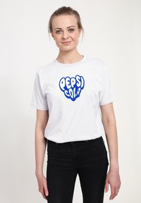 Bílý bavlněný tričko s modrým logem "PEPSI COLA" ve výrazném, zaobleném písmu. Model stojí vzpřímeně v černých džínách.