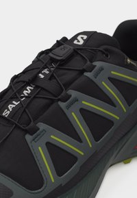 Κοντινή λήψη μαύρου και γκρι παπουτσιού για τρέξιμο σε μονοπάτι με σύστημα κορδονιών γρήγορης έλξης και κίτρινα στοιχεία στο πλάι.