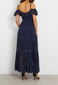 Navy blue maxi dress with off-the-shoulder ruffles, floral embroidery, and a tiered skirt, paired with black heeled sandals.