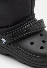 Croc noir avec une finition mate, présentant un dessus rembourré et texturé, des trous de ventilation et une sangle avec logo.