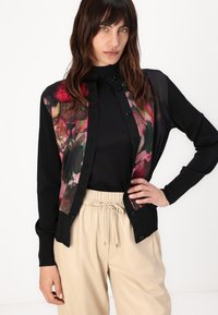 Zwarte cardigan met bloemenprint aan de voorkant, met knopen in het midden. Draagt over een zwart top met beige, trekkoordbroeken.