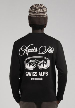 Vue arrière d'une personne portant un bonnet tricoté à motifs et un t-shirt noir imprimé de lunettes de ski, de paysages montagneux, et avec l'inscription « Après Ski, Swiss Alps Prohibited ».