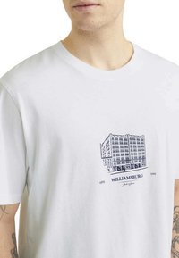 Camiseta de algodón blanca con un estampado gráfico azul de un edificio y el texto "WILLIAMSBURG", con cuello redondo y mangas cortas.