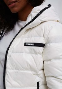 Giacca puffa bianca con cerniera nera, caratterizzata da un'etichetta nera prominente con la scritta "JACKIT". Texture liscia con sezioni trapuntate.