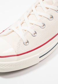 Baskets en toile beige avec lacets blancs, capuchon en caoutchouc à l'avant, rayure rouge le long de la semelle, et œillets en métal argenté. Semelle texturée blanche pour l'adhérence.