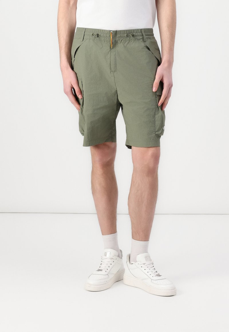 Khaki Cargo-Shorts mit elastischem Bund, seitlichen Taschen und einer lockeren Passform. Strukturiertes Material mit einem Outdoor-Design, kombiniert mit weißen Sneakers.