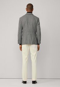 Houndstooth patterned blazer in grey and black, featuring a notch lapel and vented back, paired with cream trousers and black shoes.