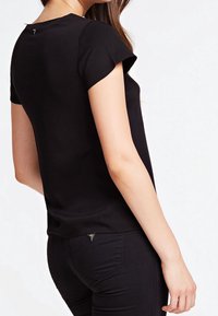Schwarzes Kurzarmtop mit rundem Ausschnitt, glattem Stoff, leichtem Glanz und einem kleinen dreieckigen Akzent auf der Rückseite nahe dem Ausschnitt.