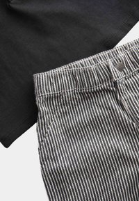 Czarny, prążkowany t-shirt, noszony na poziomych, czarno-białych spodniach; cechy to metalowy guzik i szczegóły szycia na pasie.