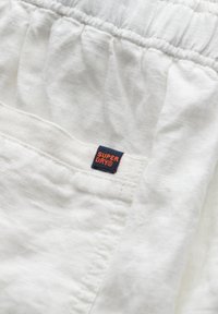 Witte katoenstof met een textuurafwerking, voorzien van een elastische tailleband en een zichtbaar "SUPERDRY" logo label in marineblauw en oranje.