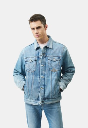 Giovane uomo con capelli corti che indossa una giacca di jeans azzurro chiaro e jeans abbinati, mani nelle tasche della giacca, sfondo neutro.
