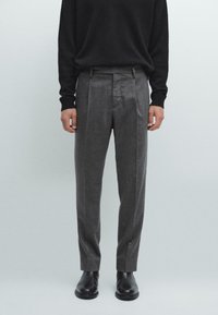 Pantaloni in lana grigi con pince e vestibilità su misura, abbinati a un maglione nero in maglia, presentati su uno sfondo neutro.