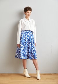 Personne debout contre un mur gris clair, vêtue d'une chemise blanche boutonnée, d'une jupe fleurie bleue et blanche, et de bottines crème sur un sol en bois.