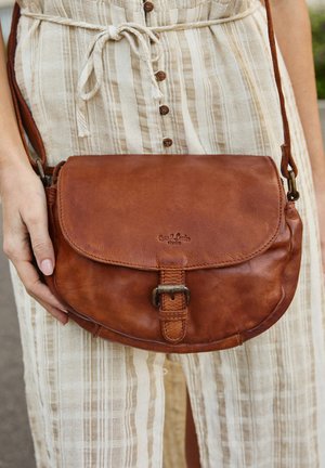 Rjava usnjena torbica preko telesa z zaklepom na preklop, podrobnostjo iz kovine ter odtisnjenim logotipom. Gladka tekstura in zaobljena oblika z nastavljivim trakom.