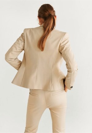 Femme aux longs cheveux roux attachés en queue de cheval, vêtue d'un blazer beige cintré et d'un pantalon assorti, debout les mains sur les hanches, de dos.