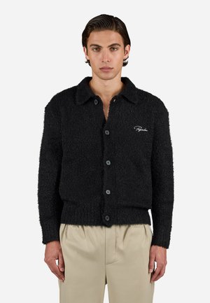 Man in een zwarte, fuzzy, knoop-up cardigan met kraag en beige broek, staande tegen een effen achtergrond.