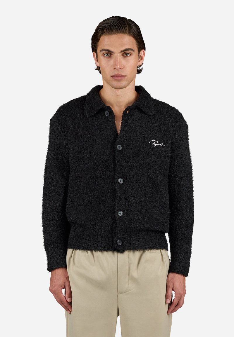 Mand iført en sort, fuzzy knappet cardigan med krave og beigefarvede bukser, der står foran en ensfarvet baggrund.