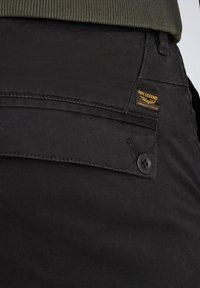 Črne hlače z gladko teksturo, sprednjim gumbom in zadrgo ter oblnik žepom z oranžno blagovno znamko na zadnji strani.