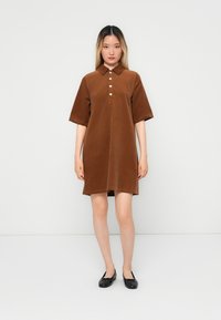 Robe en velours côtelé marron avec col, à manches courtes, dotée d'une patte de boutonnage, d'une coupe droite et d'une texture lisse. Associée à des ballerines noires.