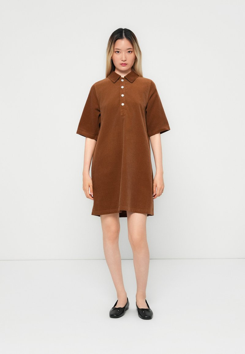 Robe en velours côtelé marron avec col, à manches courtes, dotée d'une patte de boutonnage, d'une coupe droite et d'une texture lisse. Associée à des ballerines noires.