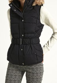 Kobieta ubrana w czarną pikowaną kamizelkę bez rękawów z paskiem, na jasnoszarym topie z długim rękawem i czarno-białych spodniakach w wzór.