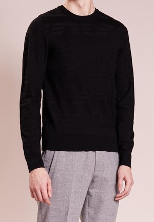 Ein Mann trägt einen schwarzen strukturierten Pullover mit langen Ärmeln und eine hellgraue, gefaltete Hose vor einem neutralen Hintergrund.
