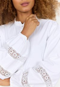 Blusa bianca con maniche voluminose, caratterizzata da un bordo in pizzo con motivi floreali. Il materiale appare morbido con una vestibilità strutturata eamp; ampia.
