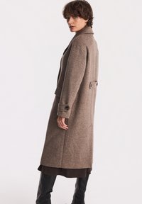 Długi brązowy płaszcz wykonany z teksturowanej tkaniny, z ząbkowanym kołnierzem, dwoma guzikami przy mankietach oraz paskiem na plecach.