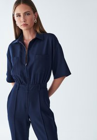 Navyblauwe jumpsuit met korte mouwen, rits aan de voorkant en twee borstzakken. Gestructureerd ontwerp met een getailleerde taille en wijde pijpen. Zijden textuur.