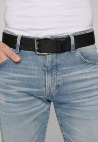 Schwarzer Leder-Gürtel mit einer silbernen Metallschnalle, ausgestattet mit mehreren Größenlöchern, getragen über hellblauen, distressed Jeans.