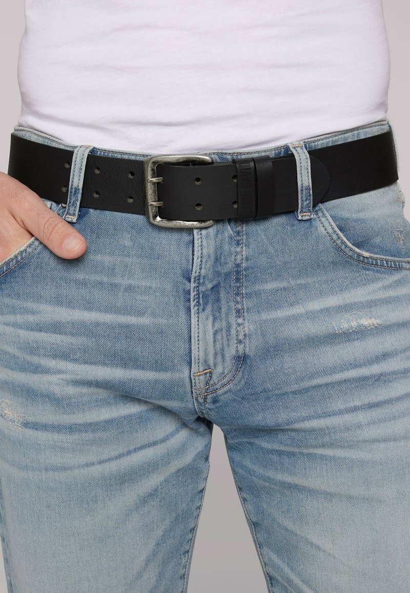 Schwarzer Leder-Gürtel mit einer silbernen Metallschnalle, ausgestattet mit mehreren Größenlöchern, getragen über hellblauen, distressed Jeans.