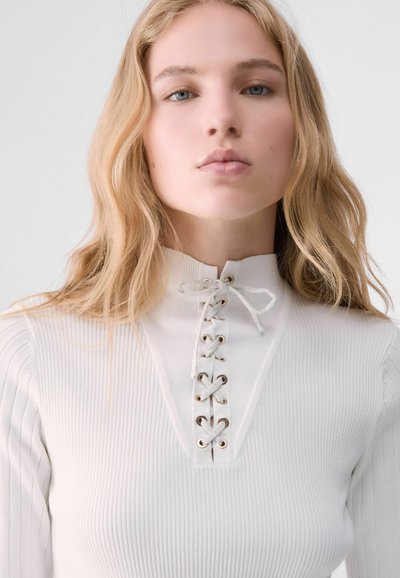 Jeune femme aux longs cheveux blonds portant un haut blanc côtelé avec un détail lacé au col montant.