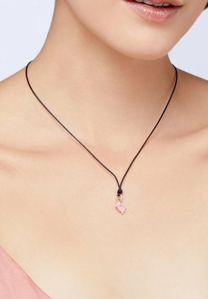 Black cord necklace with small pink dog-shaped charm worn around neck, paired with light pink top.